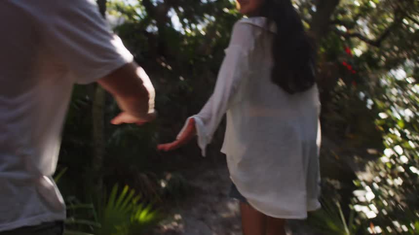 The video shows a person walking along a path through a dense lush green forest with palm fronds and other foliage the camera follows from behind as the person moves through the sundappled trail