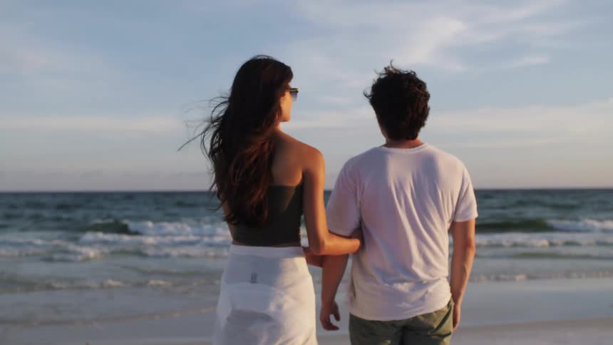 A young woman wearing sunglasses walks up to a man standing on the beach sand watching the ocean waves and lays her head on his shoulder as a light wind blows her hair at midday
