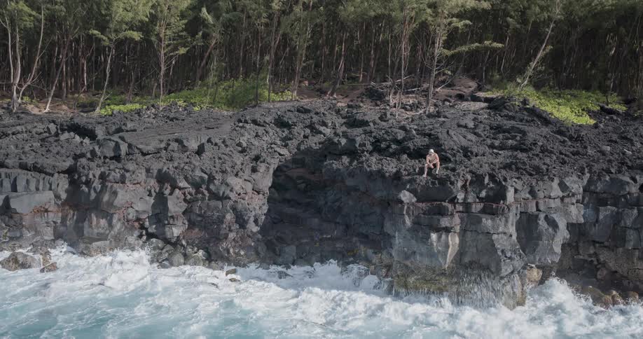 A man sits on a rocky cliff above the ocean on a partly cloudy day
