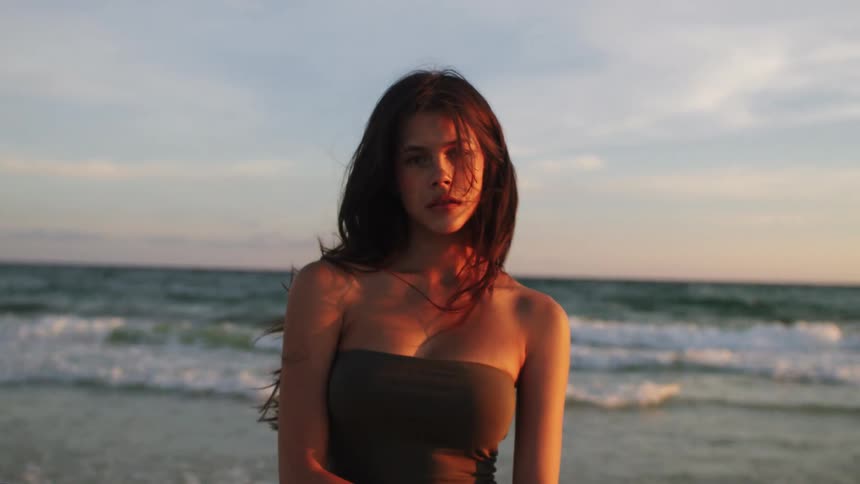 A young woman wearing a black top stands and poses on the beach as the wind blows her hair around at midday