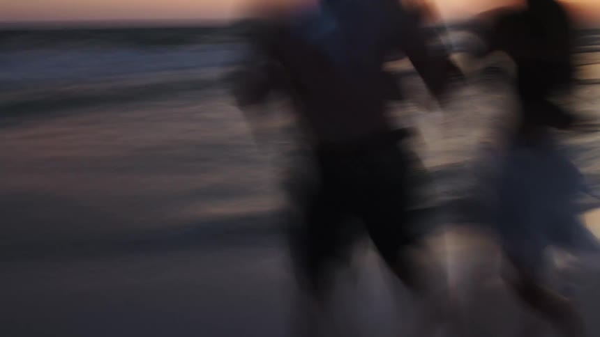 Two blurry figures of people are running along a sandy beach towards the viewer with a soft pastelcolored sunset and the ocean in the background