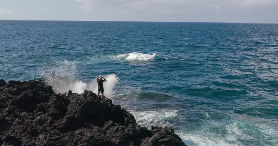 Young man standing on top of a cliff watching waves roll in and crash against the promontory