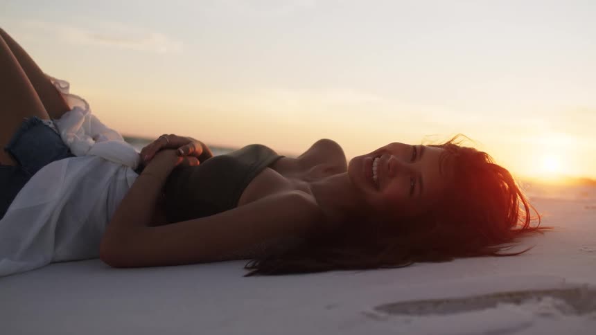 A young woman lies on the beach sand at sunset wearing a black top with a smile on her face