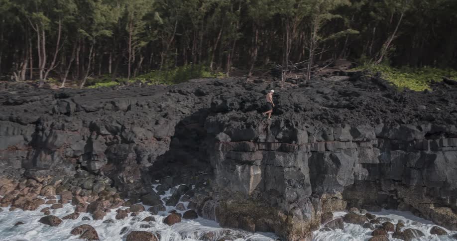 A man wearing shorts sits on the edge of a rocky cliff by the shoreline then stands up and starts walking away as ocean waves continue to crash against the rocks at midday