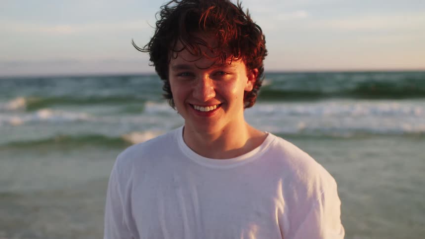 A teenage boy wearing a white tshirt stands on the beach smiling at midday