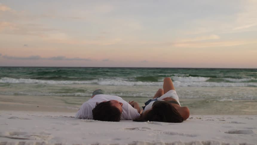 A young couple lies on the beach sand talking and looking at each other at sunset as ocean waves hit the shore
