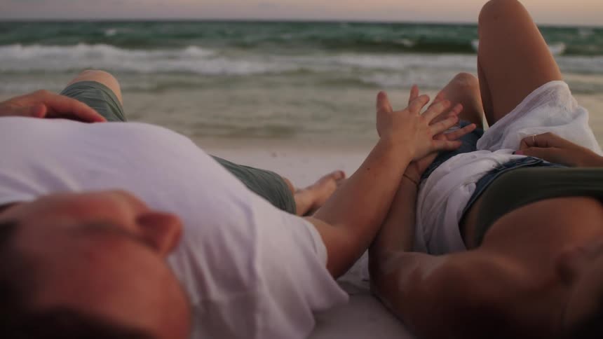 A man and a woman are lying on their backs on a sandy beach with their heads close together and their hands clasped as waves crash on the shore in front of them