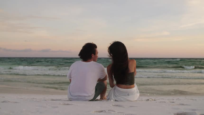 Couple sitting on white sand looking at the sea at sunset