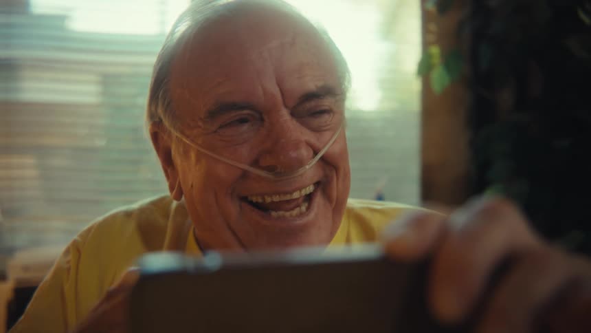 A happy smiling man wearing a yellow us army veteran tshirt and an oxygen tube is sitting in a chair and holding a smartphone raising his arm in excitement while looking at the screen