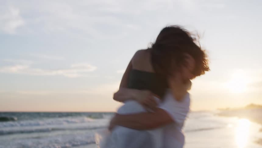 A young couple is playfully embracing on a beach at sunset with a man wearing a white tshirt picking up a woman wearing a white skirt and a dark top