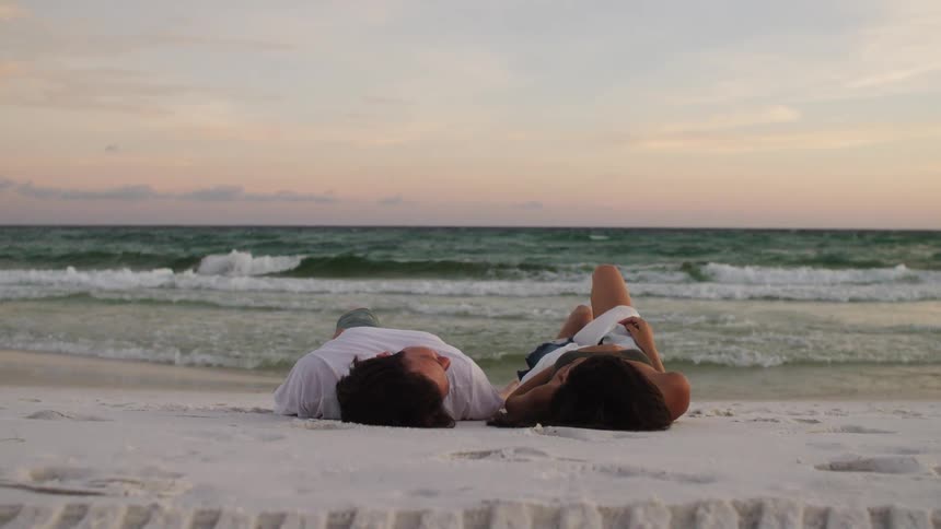 A man in a white tshirt and a woman in a white skirt are sitting on a sandy beach looking out at the waves then they both lie back and relax on the sand as the sun sets
