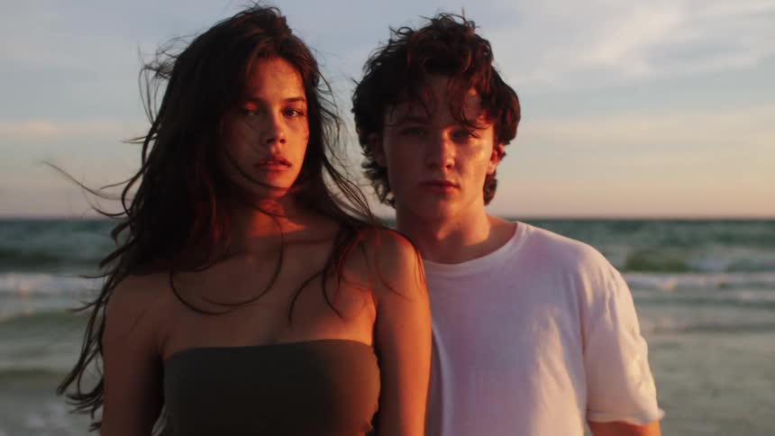 A teenage couple poses at a beach near the water line as the wind moves their hair and clothes