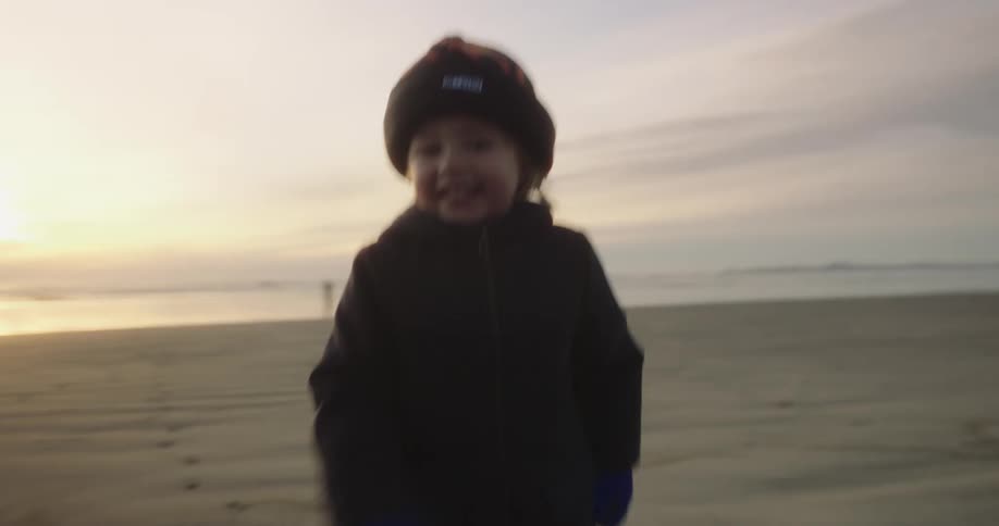 Boy wearing a coat and beanie running on the beach at sunset