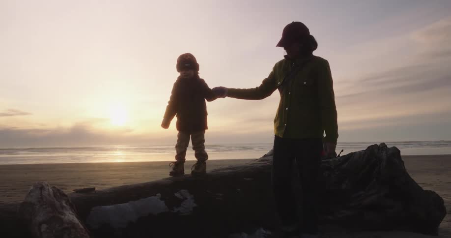A young woman and her son both wearing hats and jackets hold hands as she helps him balance while walking on a log on the beach sand at sunset