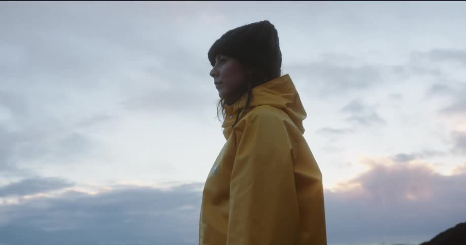 Woman wearing a yellow raincoat and black beanie looking towards the horizon at sunset