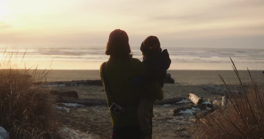 A woman carrying her toddler son stands on the beach both wearing winter jackets watching the ocean waves at sunset