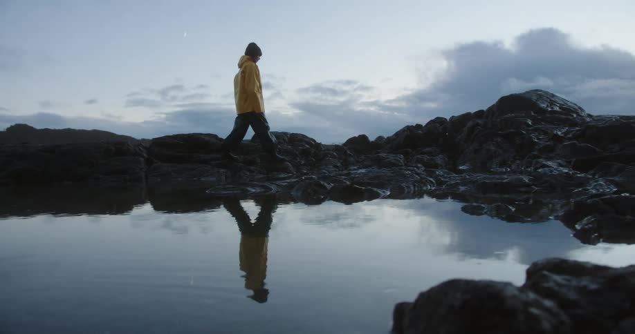 Young woman walking on rocks along the coast as clouds in the sky reflect on the water