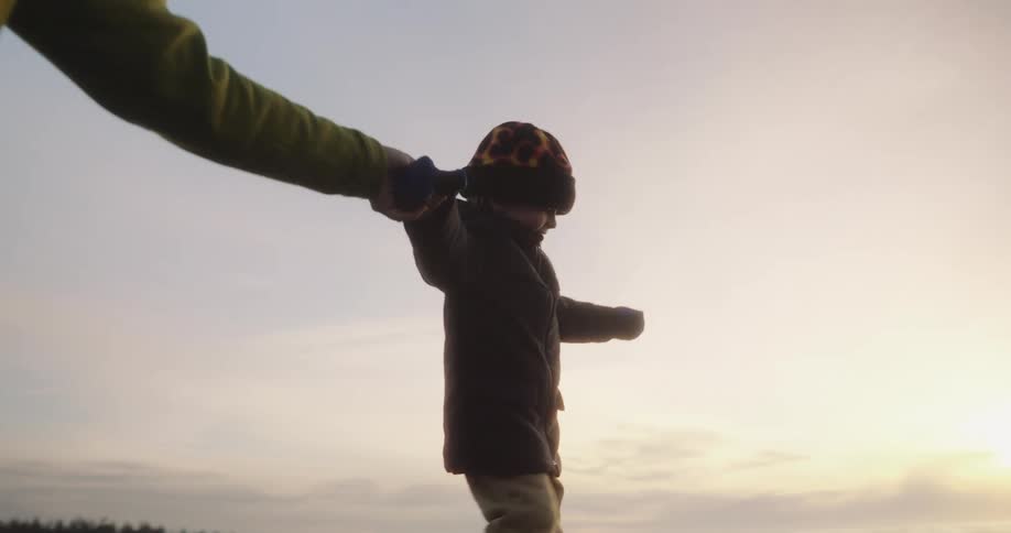 Child in a coat and beanie walks along the seashore holding an adults hand at sunset