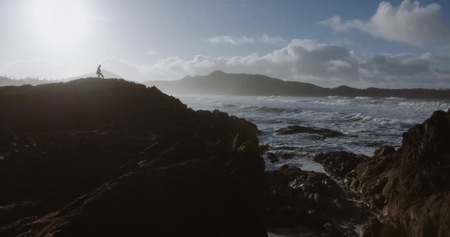 A man walking on top of a rocky island with ocean waves crashing against the rocks at midday