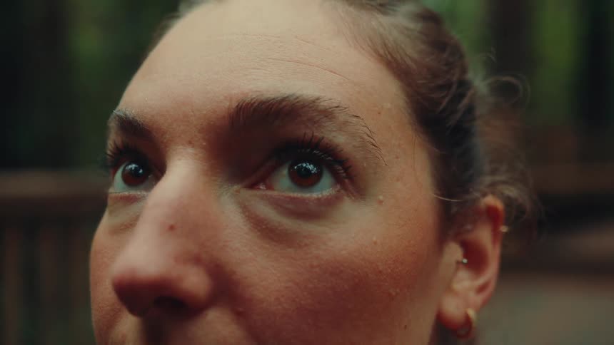 A closeup side profile of a womans face shows her eyes blinking before she look up with a nose ring multiple earrings and a blurry green forest visible in the background