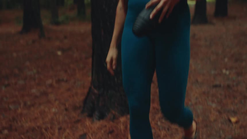 A woman with long brown hair wearing a dark blue workout outfit and a smartwatch is walking along a forest path carrying a black yoga mat stepping onto a wooden deck among the tall trees