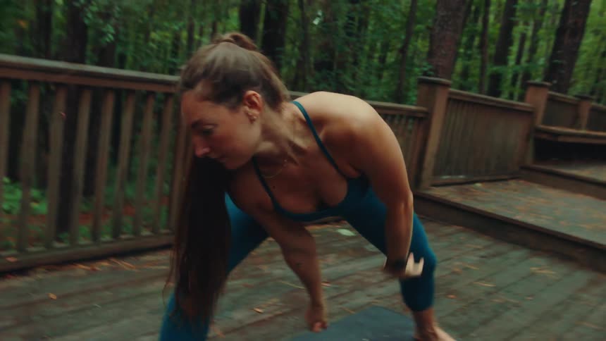 A female yogi wearing a dark blue sports bra and leggings is practicing yoga on a mat on a wooden deck in a forest transitioning from a lunge to a side lunge with her arms stretching out and up