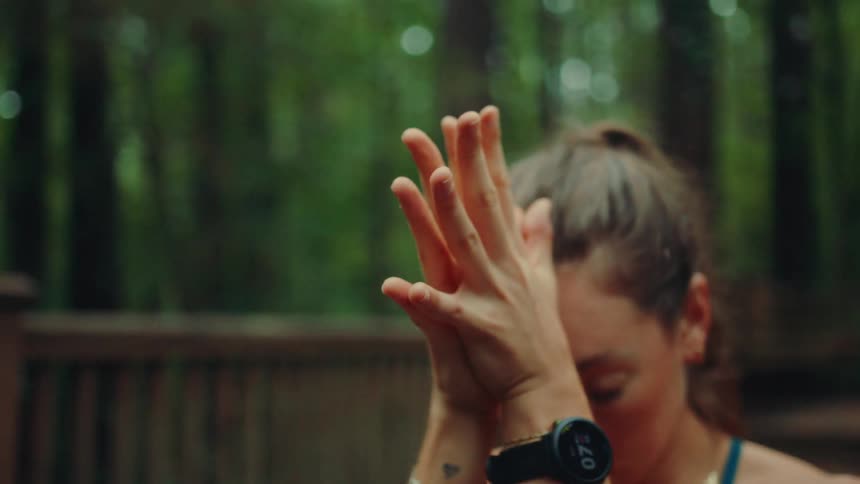 A calm woman with her hair in a ponytail is standing on a wooden deck in a forest rubbing her hands together and then bringing them to a prayer pose in front of her face