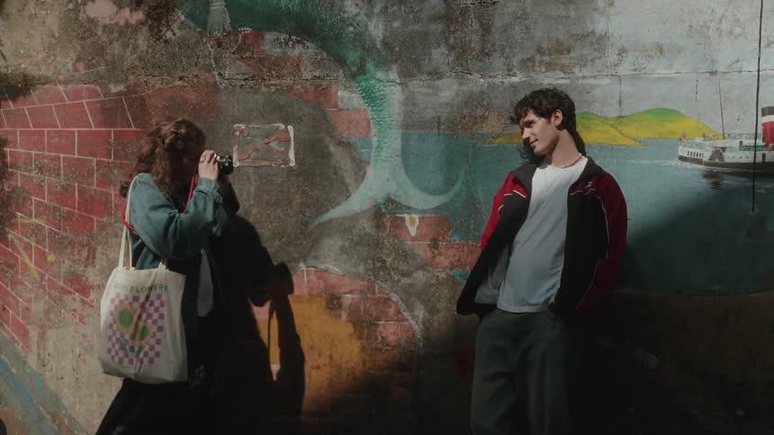 A young couple is standing in front of a brick wall with a painted mural on it as the man leans back while a woman with a tote bag raises a camera to take his picture