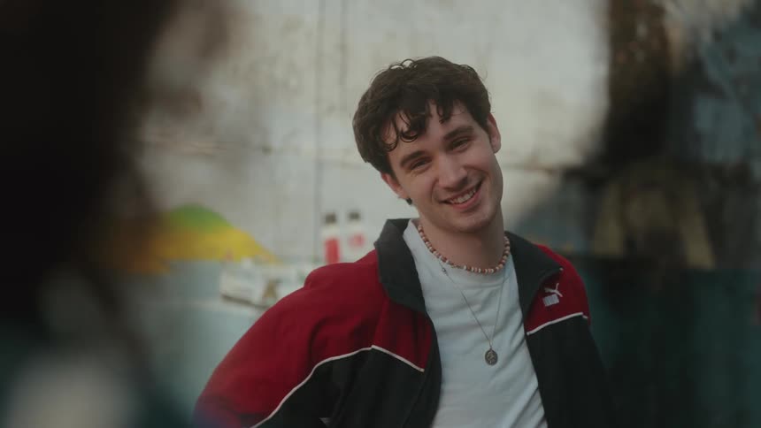 A smiling young man with messy dark hair wearing a red and black jacket and a beaded necklace is modeling for a photographer who is out of focus in the foreground with a mural on a wall in the background