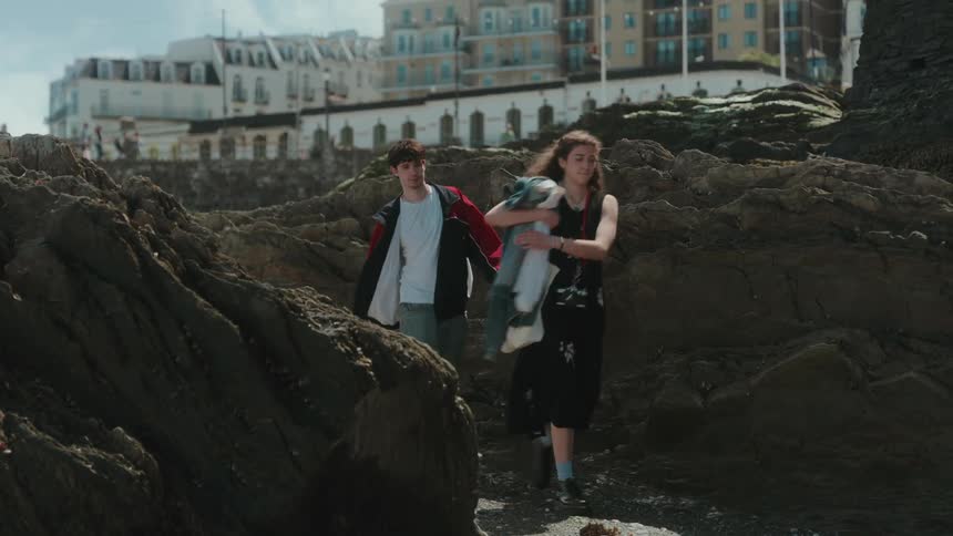 A young couple is walking along a rocky shoreline with the man wearing a red jacket and the woman wearing a black dress and carrying a gray jacket and a tote bag as white buildings are seen on a cliff in the background
