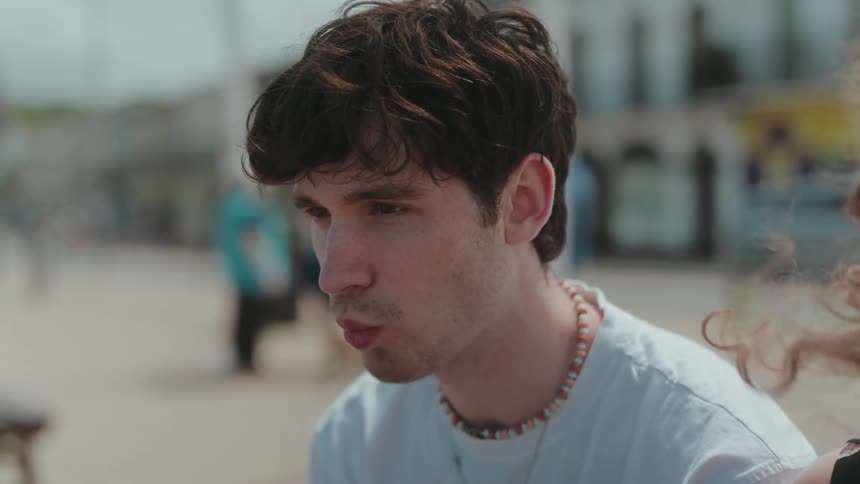 A man with messy dark hair wearing a lightcolored tshirt and a beaded necklace is seen from a side profile eating an ice cream cone on a sunny day