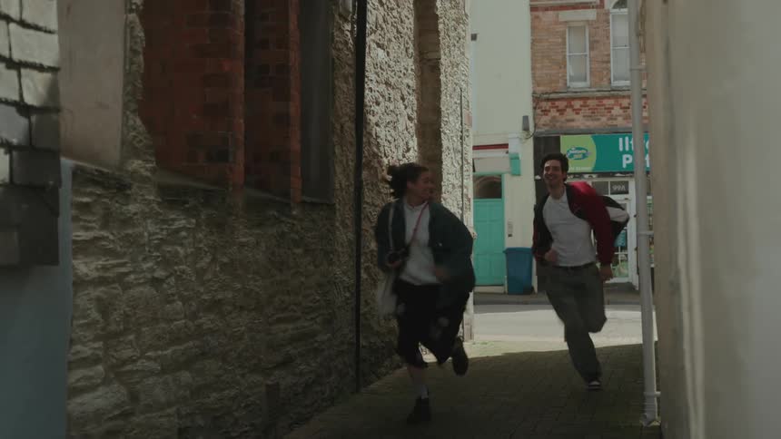 A young woman and man run through a narrow alley toward the street