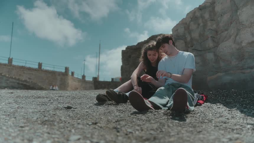Couple sitting on gravel ground near a rocky wall under a blue sky