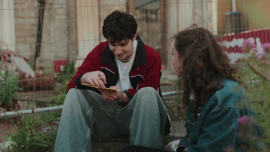 Two teenagers sit on a lawn both laughing as the boy writes in a notebook while looking at the girl both laughing at midday