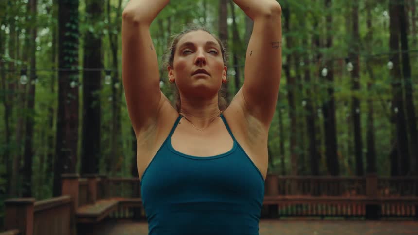 A calm woman in a blue tank top stands on a wooden deck in a forest slowly raising her arms bringing her hands together into a prayer pose and closing her eyes