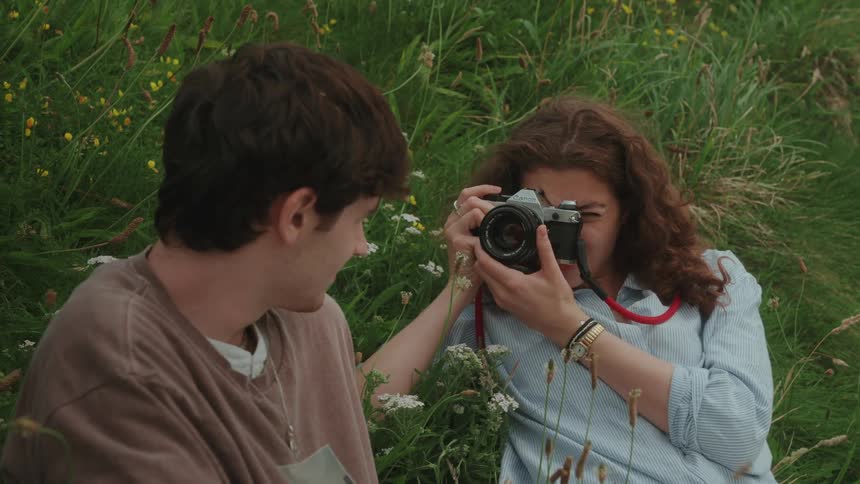 A female in a blue shirt takes a photo of someone sitting beside them in tall grass