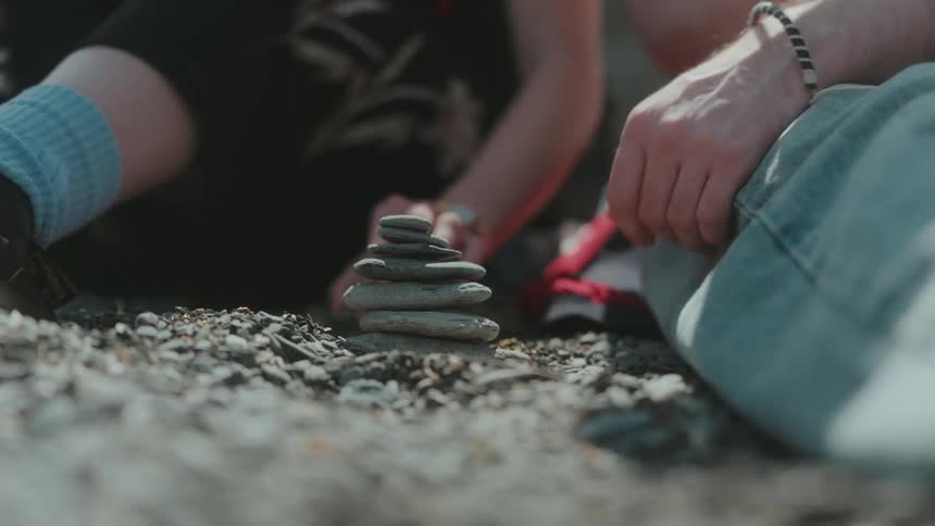 Two people seated on the ground under the sun stacking small rocks one on top of another
