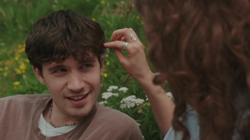 A man is lying in a field of grass with white flowers and a woman is sitting next to him talking to him and putting a flower behind his ear