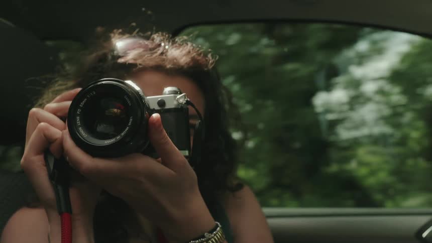 A teenager is holding a silver vintage film camera up to their eye in a driving car