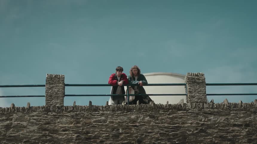 A man and a woman leaning onto a railing side by side as they talk with the woman pointing things out below them
