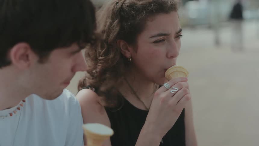 A young man and woman sit on a bench on the sidewalk in the city at midday enjoying ice cream cones