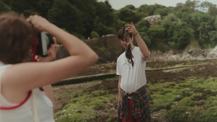 Woman photographing a man holding seaweed in a coastal setting with rocks and moss
