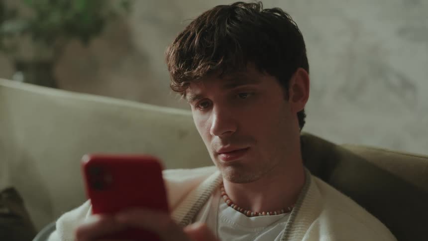 A man with short brown hair is sitting in a living room wearing a white cardigan over a tshirt and a beaded necklace as he looks at his red smartphone and runs a hand through his hair