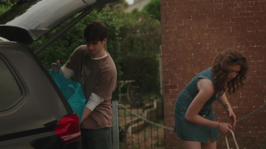 A young man with short brown hair and a young woman with curly hair are packing the trunk of a car both placing suitcases and duffel bags inside