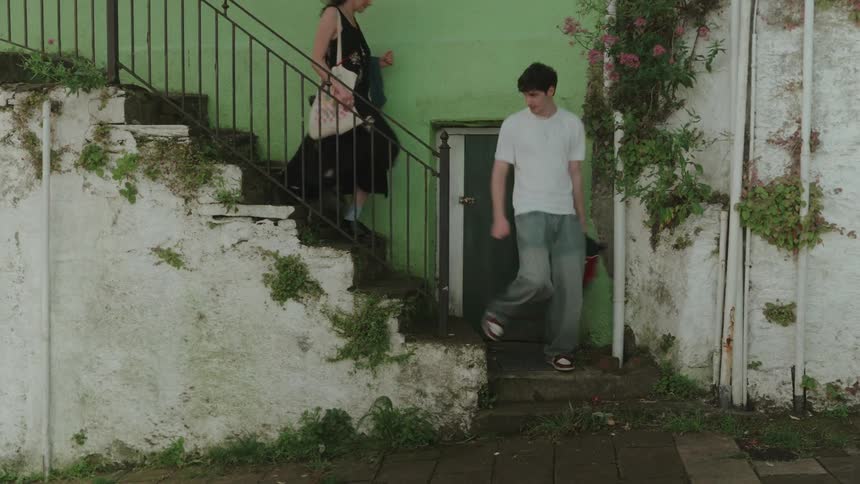 A man in a white tshirt and a woman in a dark dress are walking down a staircase with a black metal railing with a light green wall and a green door behind them and moss and vines growing on the white stone walls
