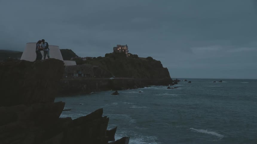 A man and woman are standing on the edge of a rocky cliff overlooking the ocean talking to each other as a small building is seen in the distance and waves crash on the rocks