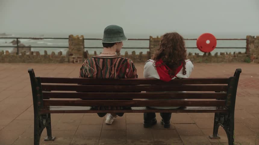 A man and woman sit on a bench by the shore