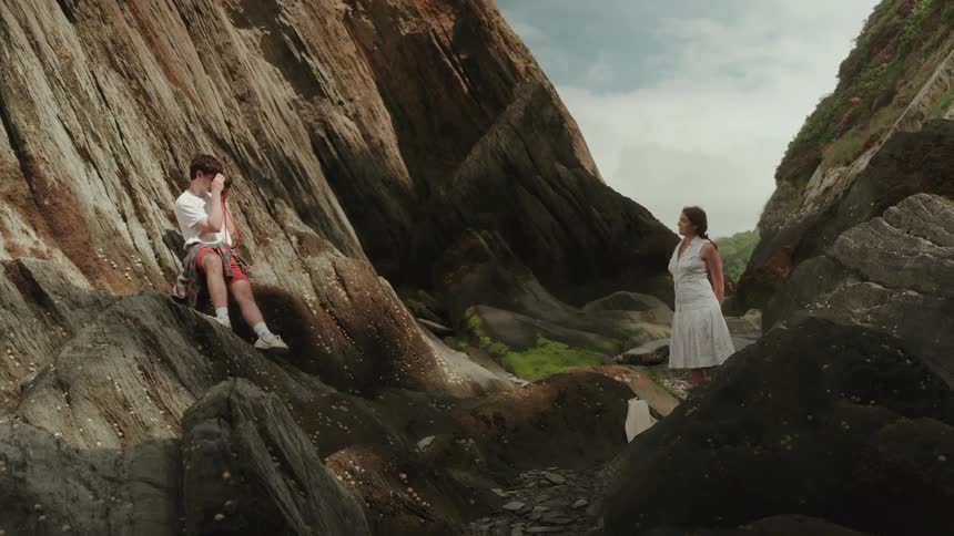 Man sitting on rocks photographs a woman in a white dress standing lower among rocky formations