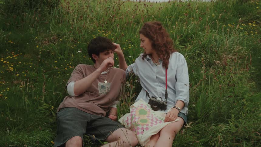 Man and woman sitting on the grass interact affectionately with a camera hanging around her neck