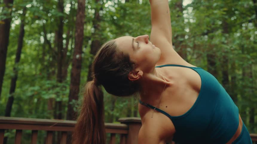 A female yogi wearing a dark blue sports bra and leggings is practicing yoga on a wooden deck in a forest transitioning from a lunge to a side bend with her arms stretching out and up